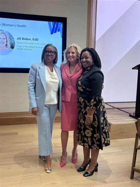 Three women standing together smiling at a professional event with presentation screen in background